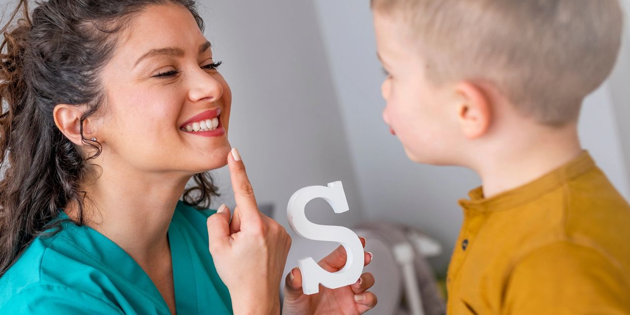 Speech therapist holds up a letter S and helps boy with stutter.  Child shapes mouth to make sound