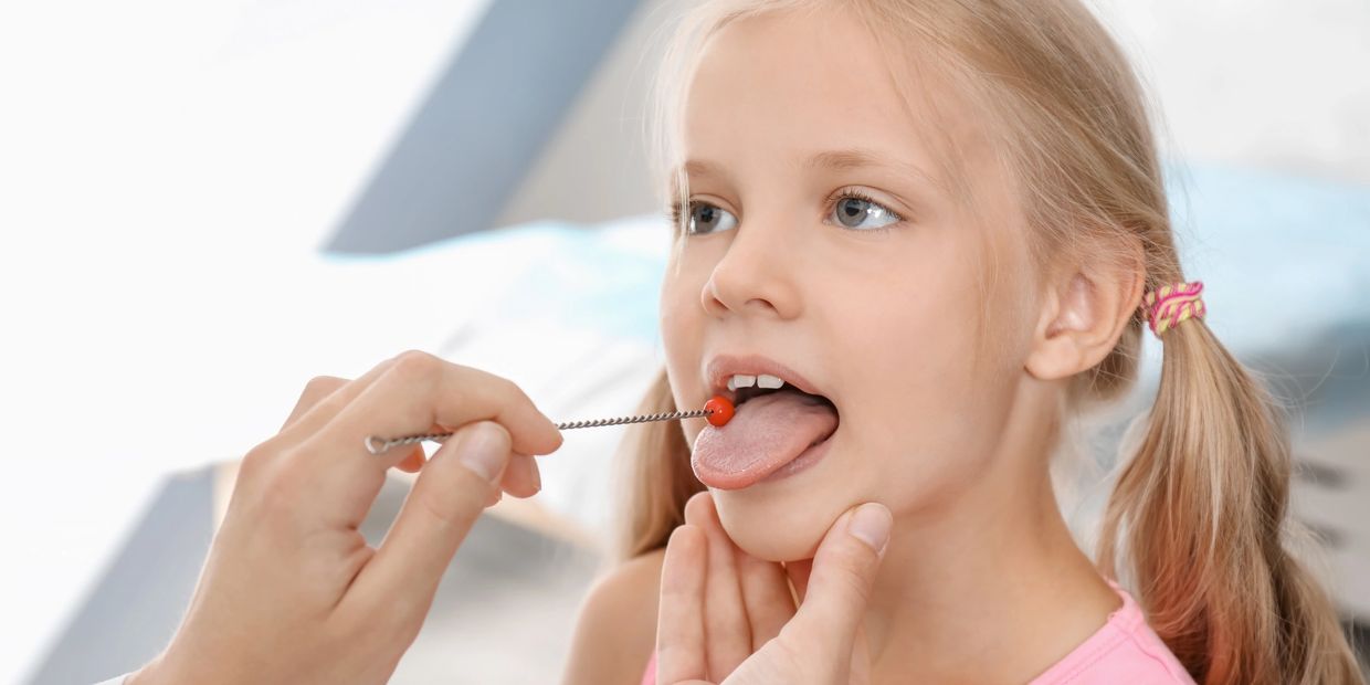 Young girl with blond hair sticks tongue out. Speech-language pathologist places long tool on tongue