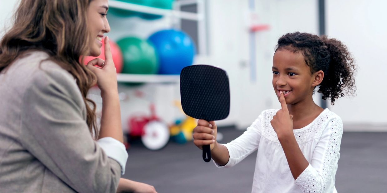 Speech language therapist sits with girl. Child holds mirror an touches her teeth with her finger.