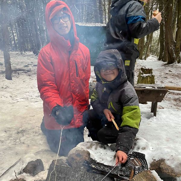 Two boys roasting hot dogs over a campfire in a snowy forest.
