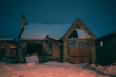 A snow-covered, abandoned house at night with boarded windows.