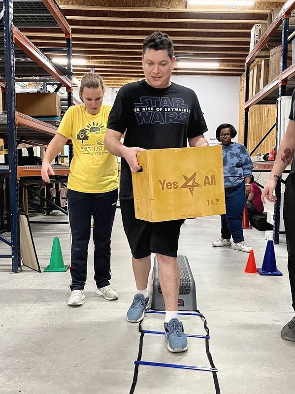 Adult male carrying a box while doing the agility ladder with two other athletes looking on.