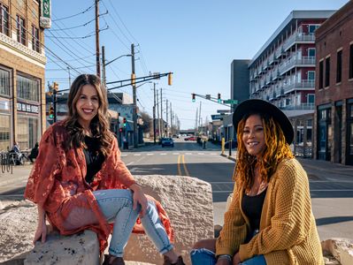 2 hairstylists sitting on rocks in a city setting and smiling at the camera