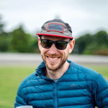 Smiling man wearing sunglasses, a cap, and a blue jacket outdoors.
