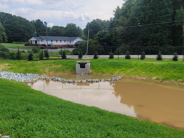 A small muddy pond with a concrete drainage structure and greenery around it.