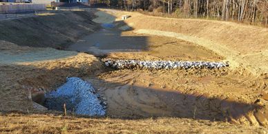 A dry, grassy drainage ditch with rock barriers and a forest in the background.