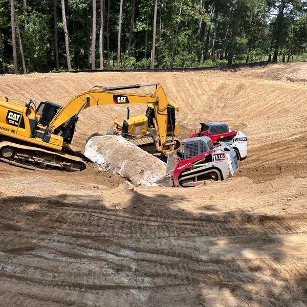 Excavators and mini skid steers working on a large dirt excavation site surrounded by trees.