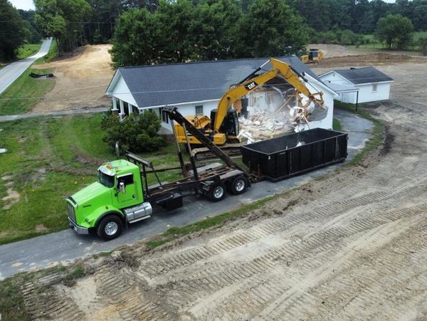Excavator demolishing a house near a green truck with a dumpster for debris.