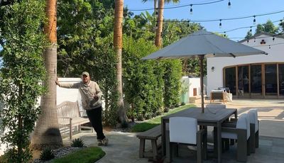 A man leaning on a palm tree in a sunny backyard with outdoor dining furniture.