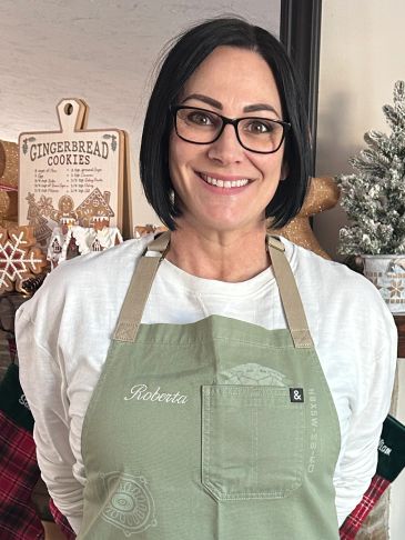 Woman smiling in glasses and green apron with holiday decor behind.
