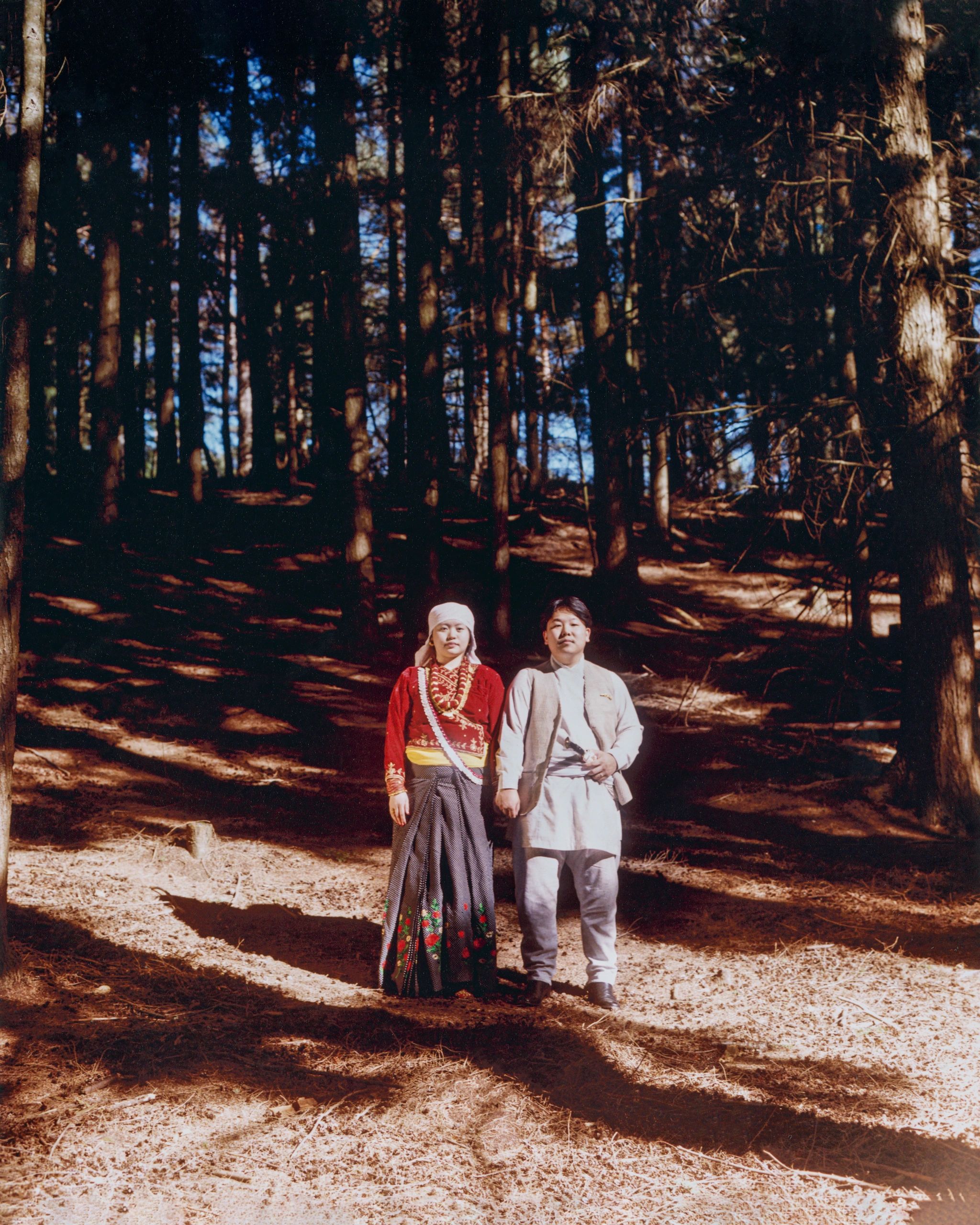 A couple in traditional attire stands in a sunlit forest clearing.