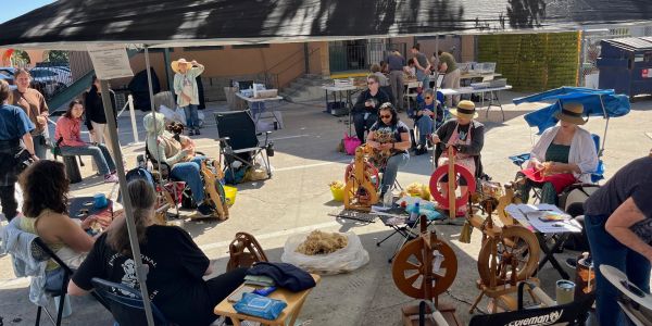 People gathered outdoors spinning yarn using spinning wheels under a tent.