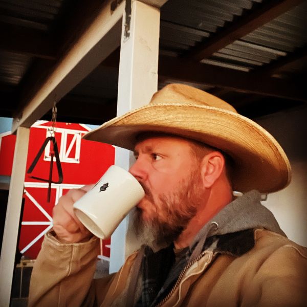 Man in a cowboy hat drinking from a white mug on a porch.