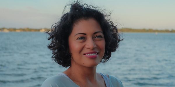 A smiling woman with curly hair stands by the water during sunset.