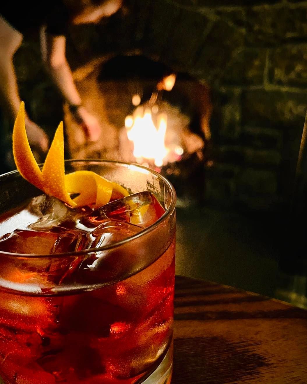 A cocktail with an orange twist near a fireplace.