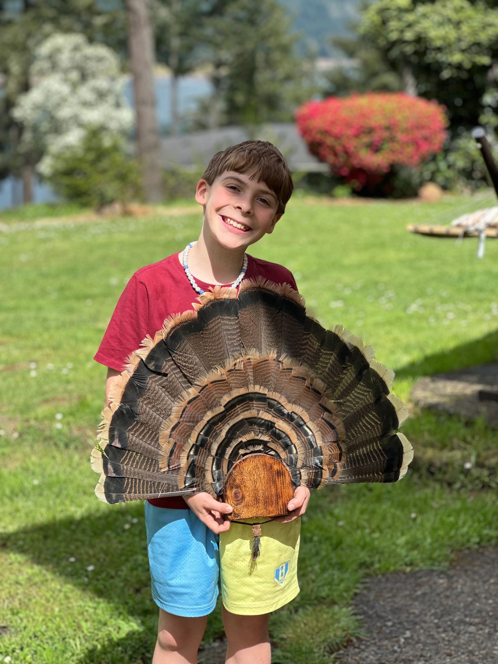 A smiling boy holding a large feather fan outdoors.