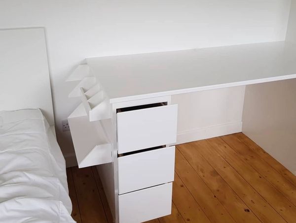 A sleek white desk with three drawers and a unique side shelf next to a bed.