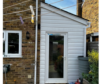 Small white garden shed with a glass door beside a brick house under a clear sky.
