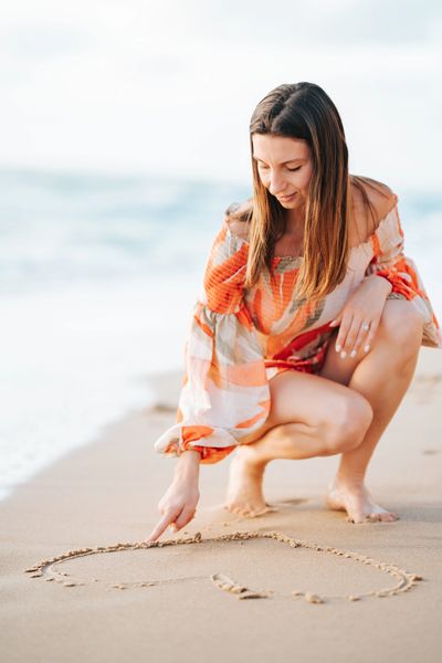 a woman at the beach in a summery outfit, drawing a heart into the sand, feeling at ease