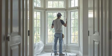 A man stands on a ladder painting window frames in a bright room.