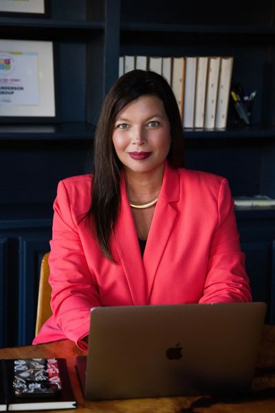 Confident woman in a coral blazer working on a laptop at a wooden desk.