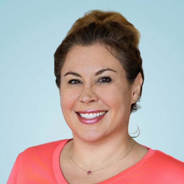 Smiling woman wearing a coral shirt with a necklace against a light blue background.