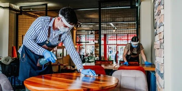 Two workers disinfecting tables in a restaurant wearing masks and gloves.