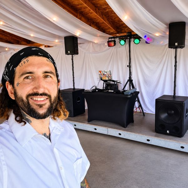 Man with bandana takes selfie in front of DJ setup on small stage.