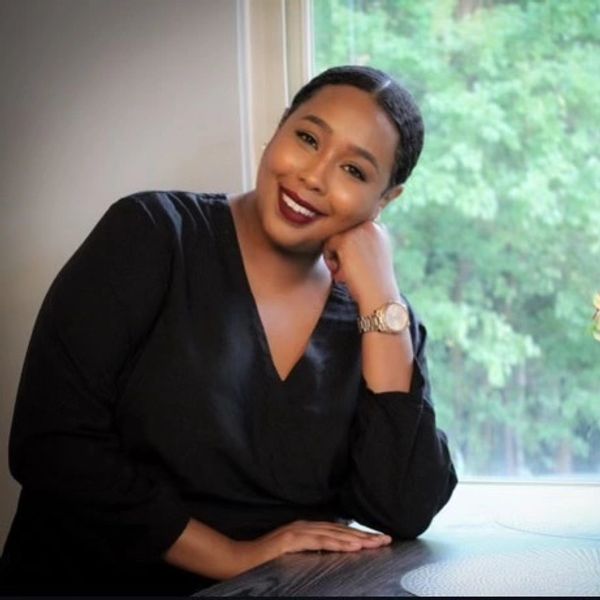 Smiling woman in black dress sitting by a window with greenery outside.