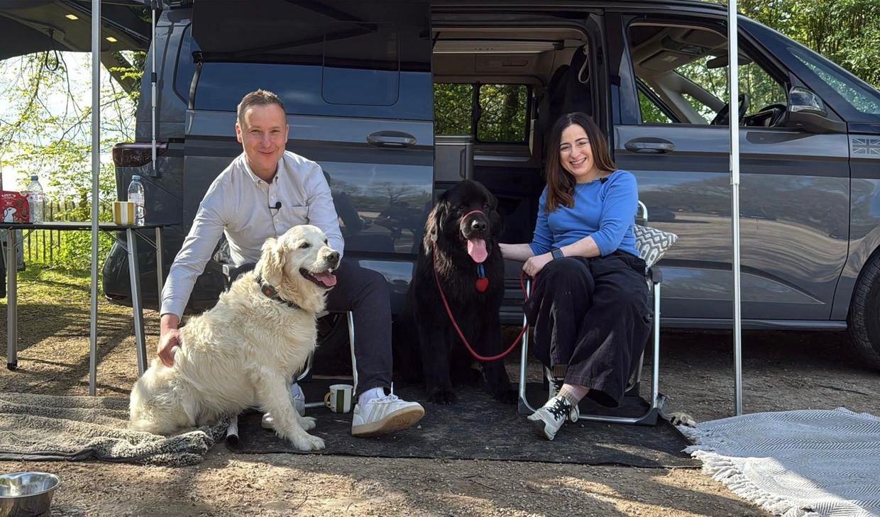 A happy couple sits with two dogs by a camper van in a sunny outdoor setting.