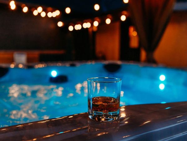 Whiskey glass on the edge of a glowing blue hot tub with warm string lights.