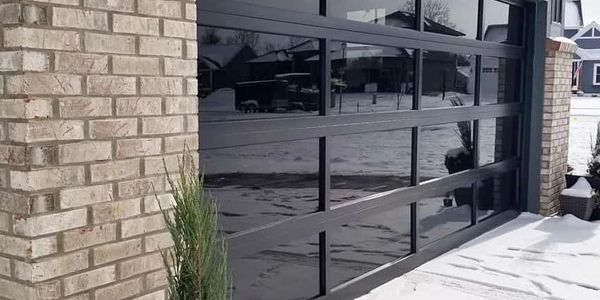 Modern black garage door with snow-covered driveway and plants in pots.
