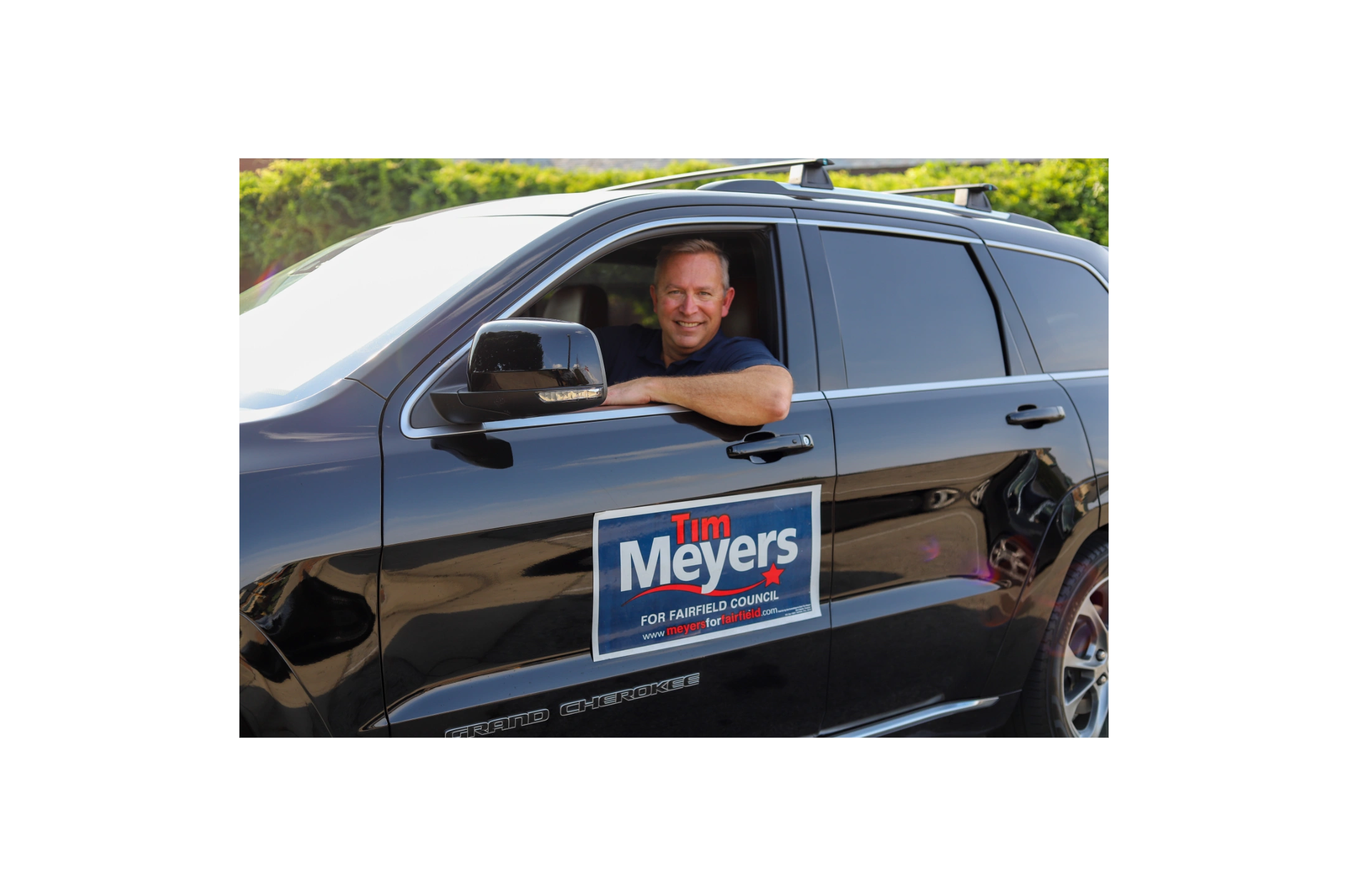 Man smiling from driver's seat of a black SUV with campaign sign.