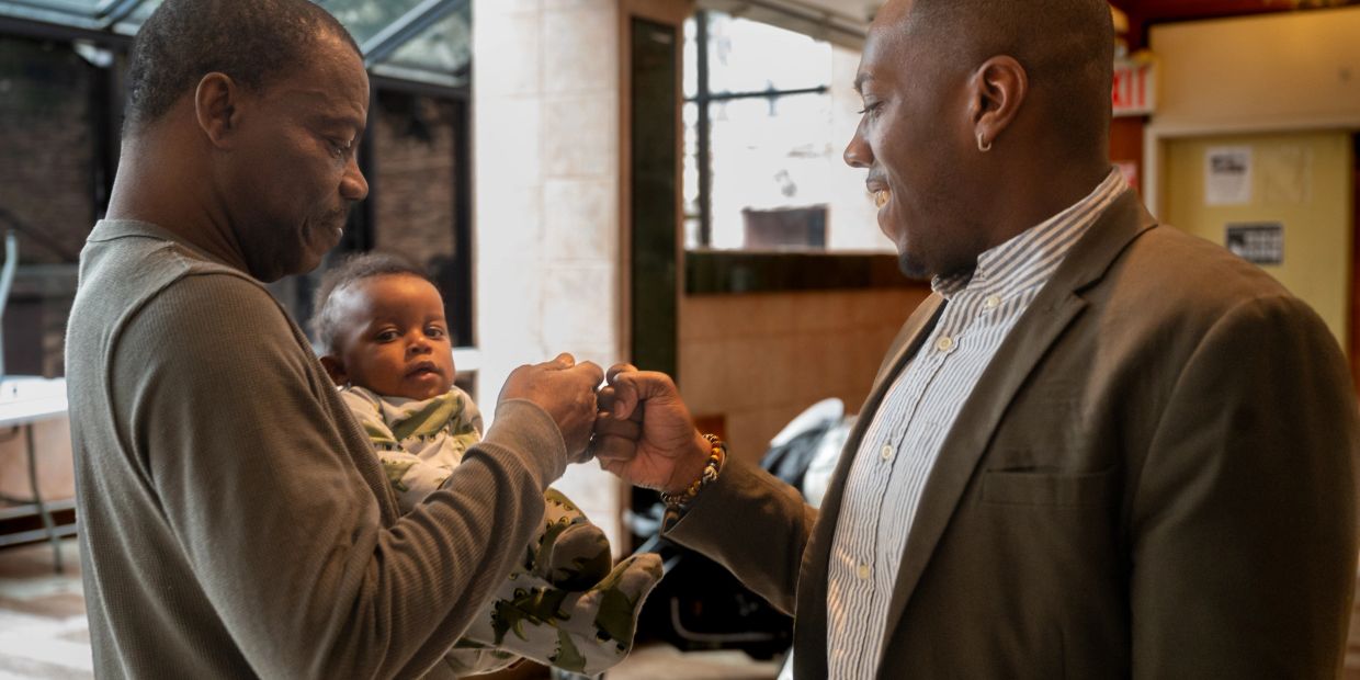 Two men fist bump while one holds a baby indoors.