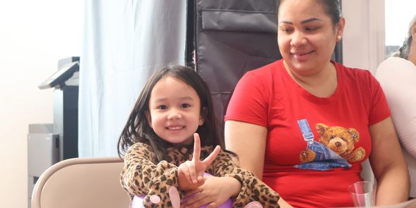A smiling child showing a peace sign beside a woman in a red shirt with a teddy bear design.