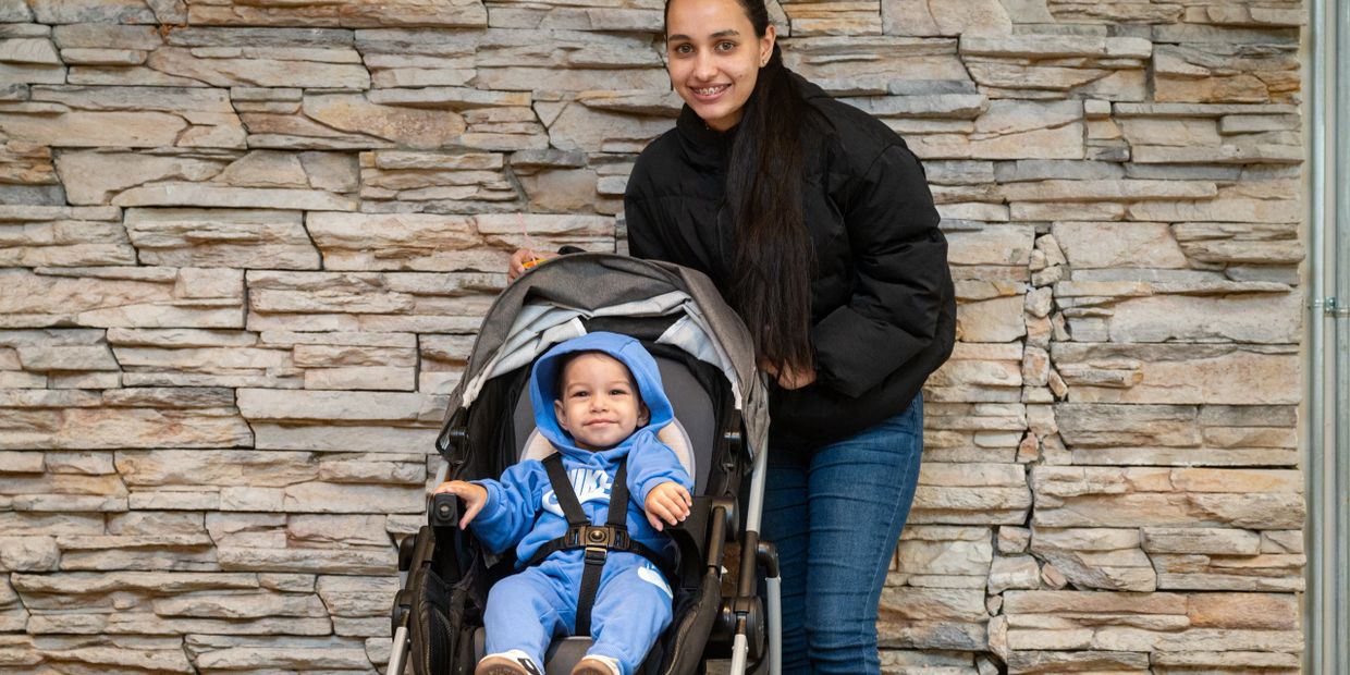 Smiling woman stands behind a baby in a stroller against a stone wall.