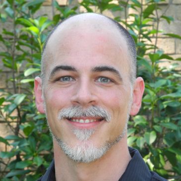 Smiling man with a trimmed beard and bald head outdoors.