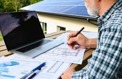 Man working on architectural plans outside near solar panels with laptop and documents.