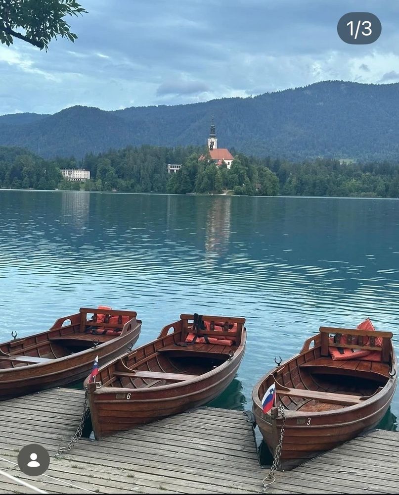 Three wooden boats docked on a serene lake with a church on an island in the background.