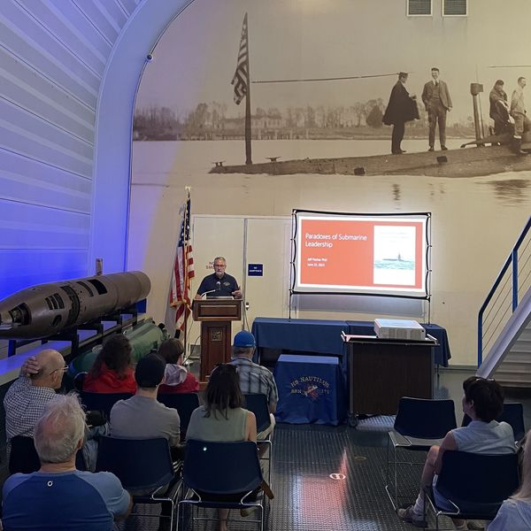 A speaker presents 'Paradoxes of Submarine Leadership' to an audience in a submarine-themed room.