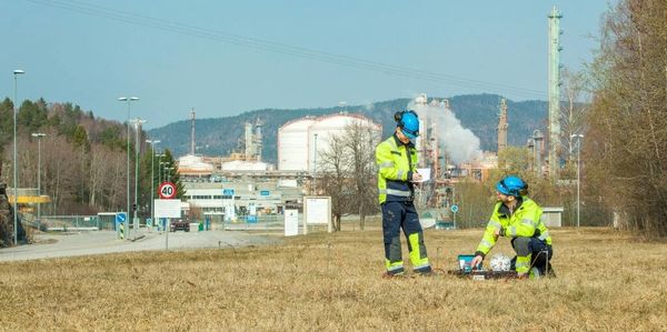 Two workers in safety gear inspecting equipment near an industrial plant.