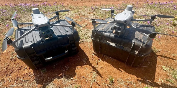 Two drones resting on black protective cases outdoors on dry ground with purple flowers.