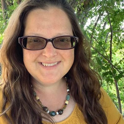 Woman smiling outdoors wearing glasses and a beaded necklace.