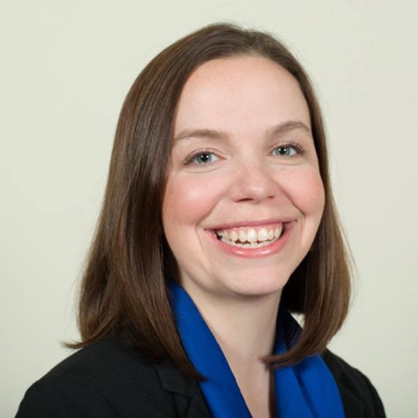 Smiling woman with brown hair wearing a black blazer and blue scarf.