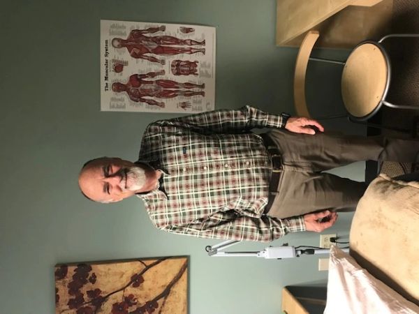 Elderly man standing in a medical or therapy room with anatomical poster.