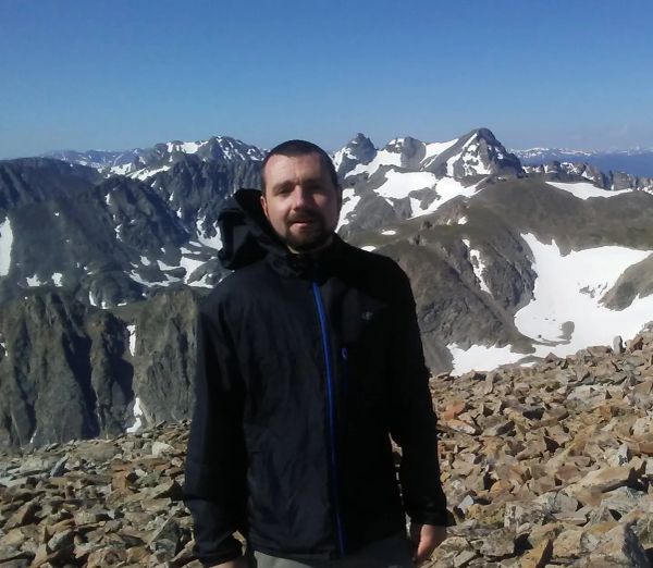 Man in black jacket standing in front of snowy mountain peaks.