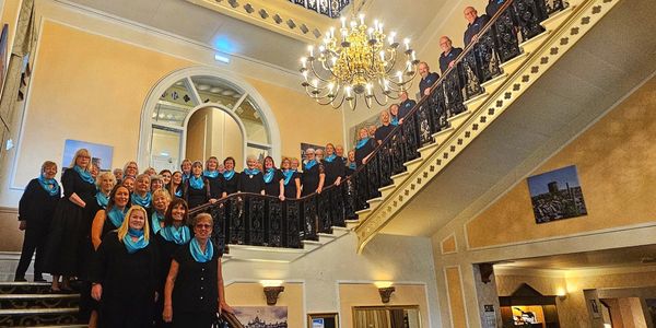 A large choir group poses on an ornate staircase under a chandelier.