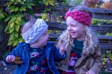 Two young girls wearing knitted headbands share a joyful moment outdoors.