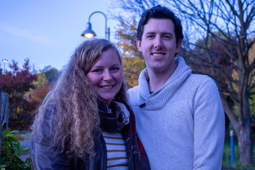 A smiling couple posing outdoors in autumn with trees and a lamp post.