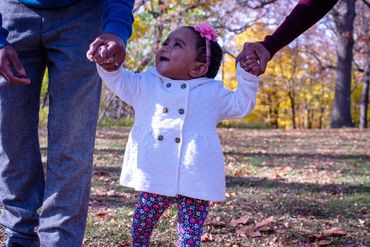 A toddler holds hands with two adults while learning to walk outdoors.